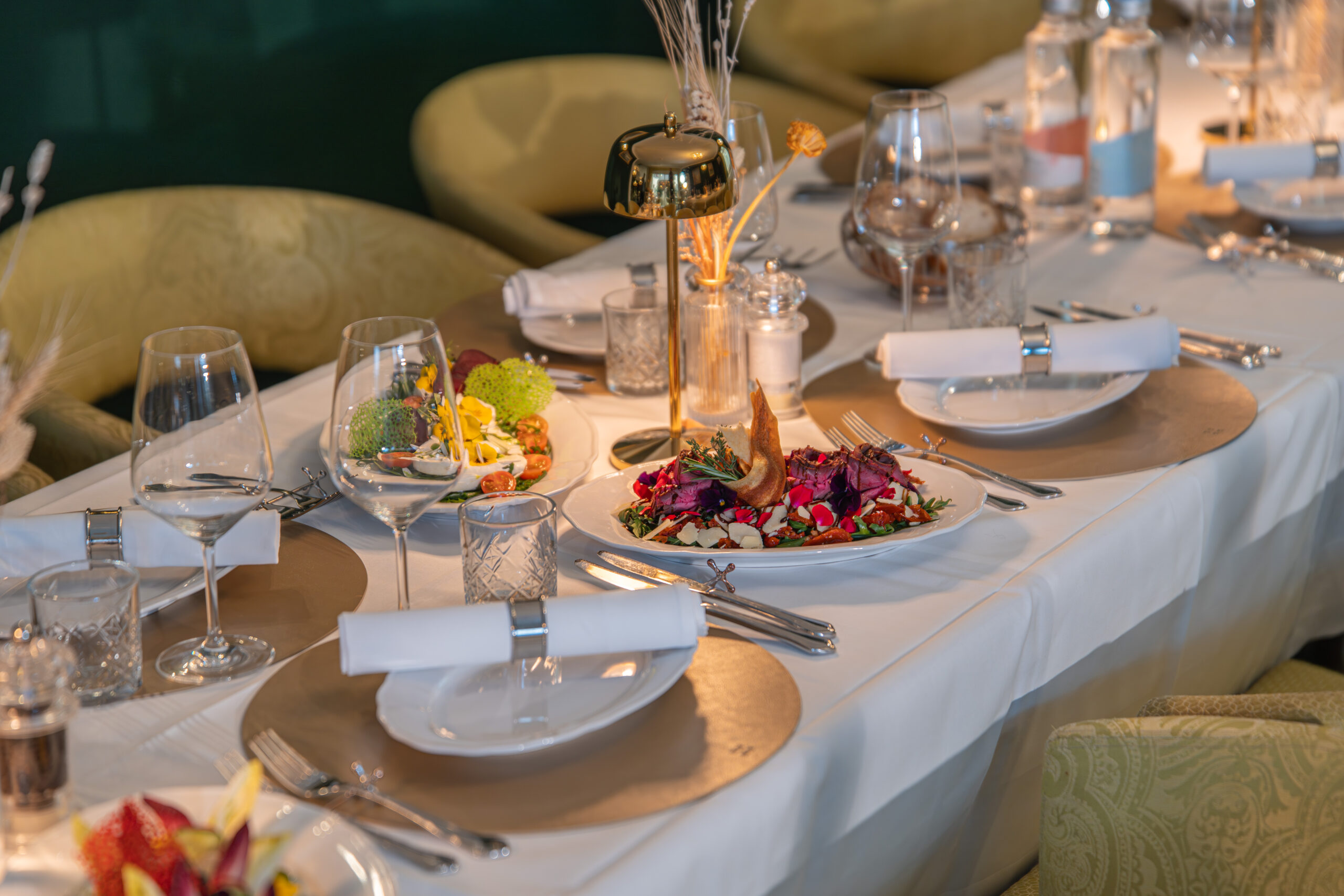 Weiss gedeckte Tafel mit goldener Lampe in der Mitte. Goldene Tischsets mit Tellern und Servietten. Bunt garnierte Speisen auf dem Tisch verteilt